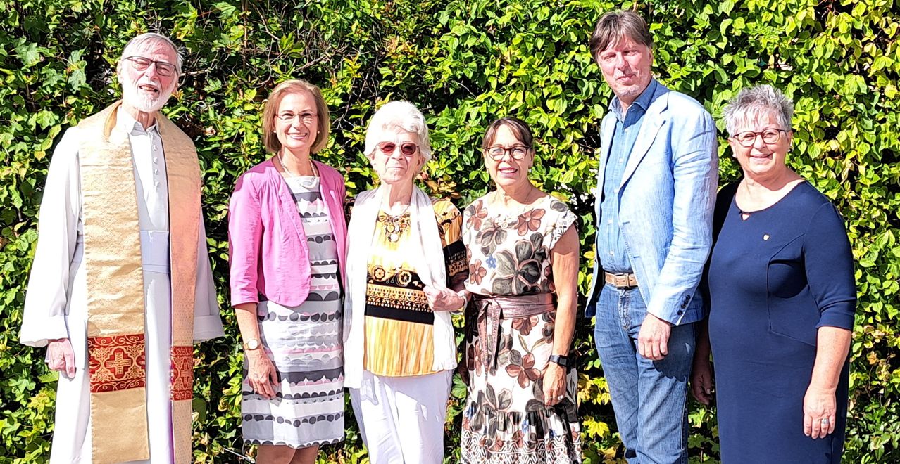 Four people stand in front of a bush, smiling for a photo. The woman on the left wears a pink jacket. The woman in the middle has white hair and wears a yellow top. The woman on the right wears glasses and a floral dress. The man on the right wears a blue shirt and jeans.