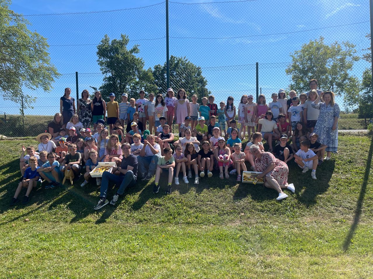 Eine große Gruppe von Kindern und Erwachsenen posiert für ein Foto auf einem Grasfeld. Einige halten Snacks, andere tragen Hüte. Bäume und ein Zaun sind im Hintergrund.