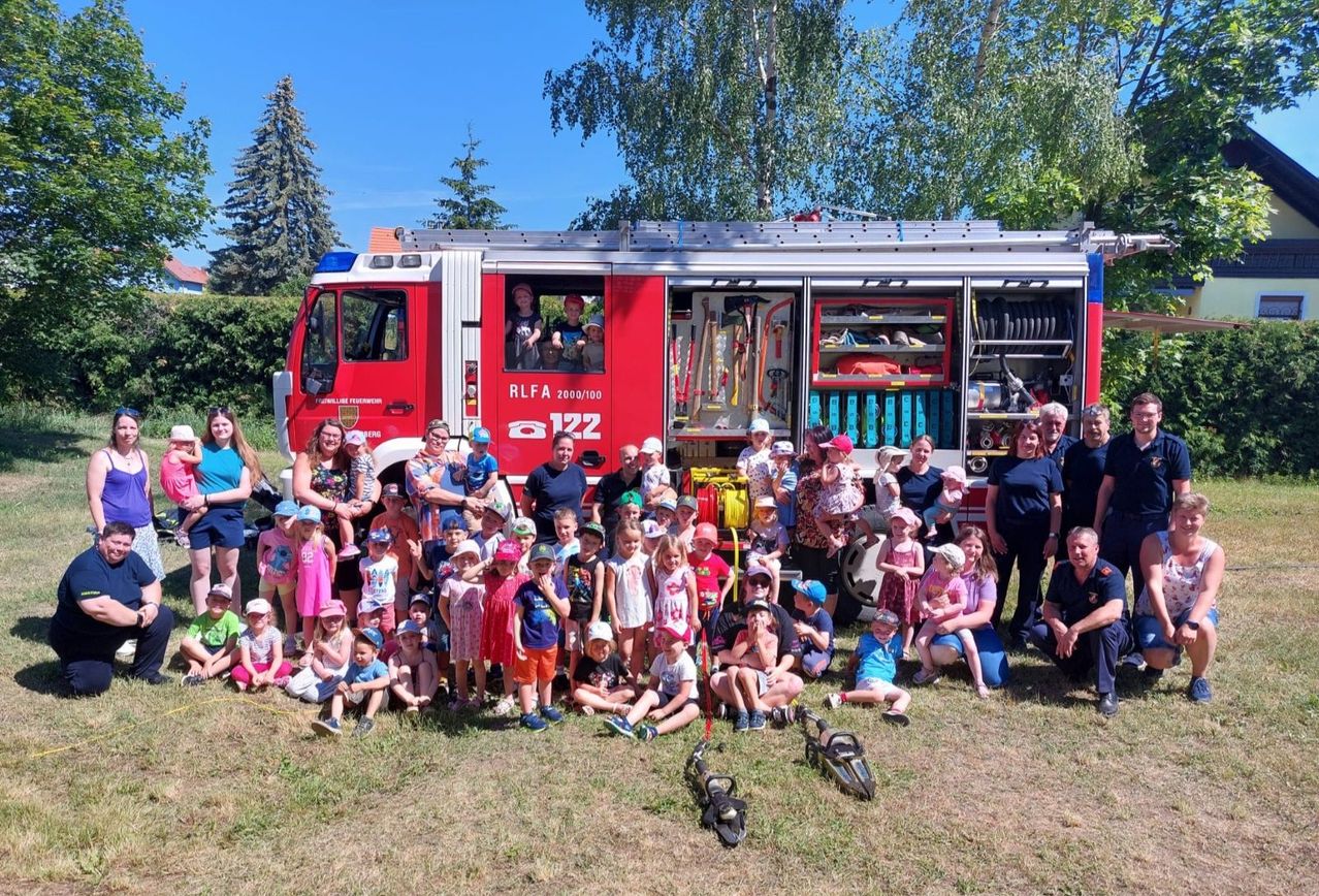 Eine Gruppe von Kindern und Erwachsenen posiert vor einem roten Feuerwehrwagen mit Personen im Inneren. Die Gruppe steht auf Gras in der Nähe einer Hecke.