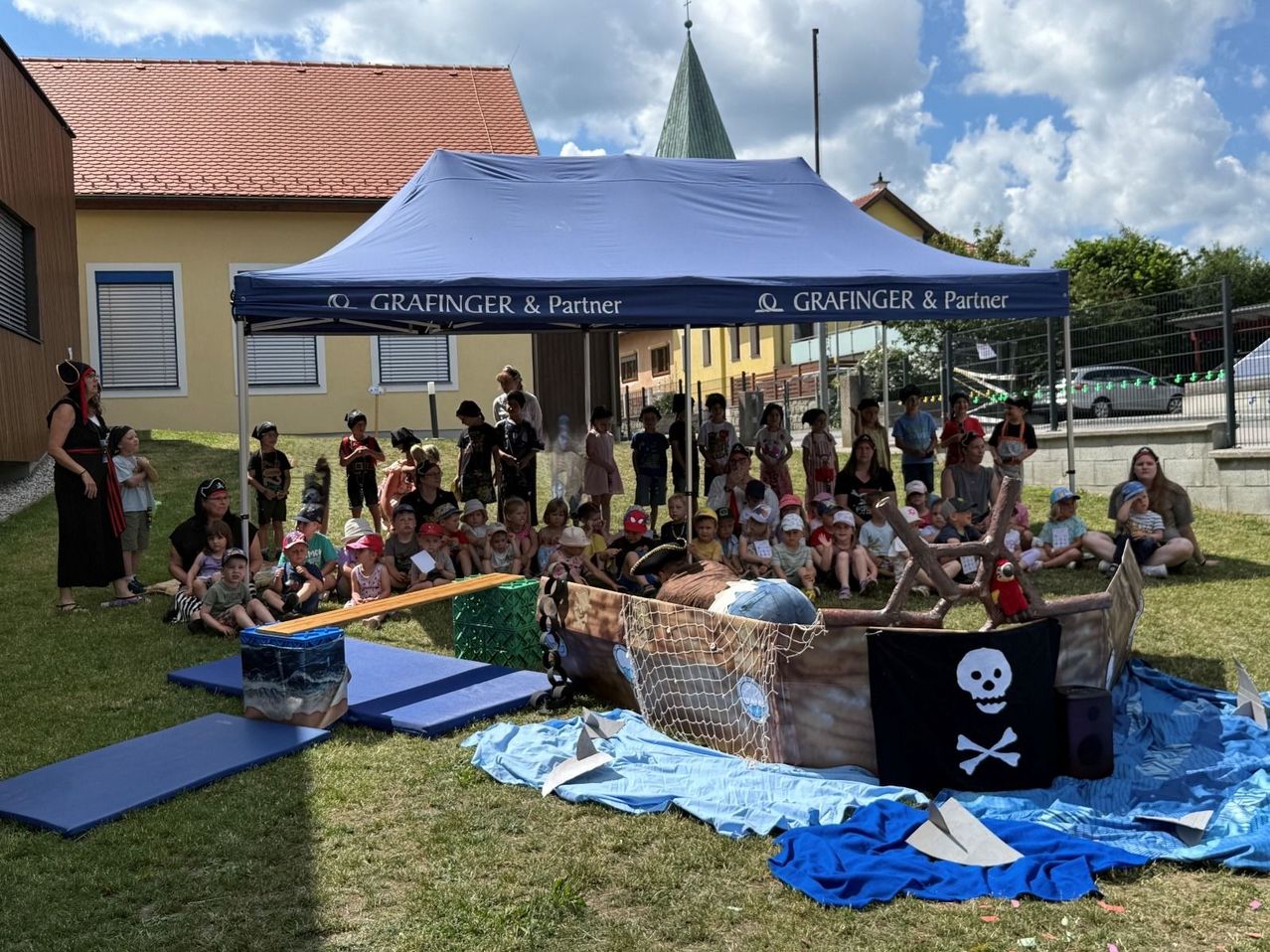 Eine Gruppe von Kindern in Piratenhüten versammelt sich um ein Piratenschiff unter einem Zelt mit der Aufschrift Grafinger & Partner. Einige sitzen auf Bänken, während andere in der Nähe des Schiffes auf einem Rasen stehen.