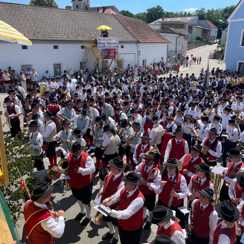 Eine große Gruppe von Musikern in traditioneller Kleidung tritt im Freien vor einer Menge auf, mit Gebäuden und Grünpflanzen im Hintergrund.