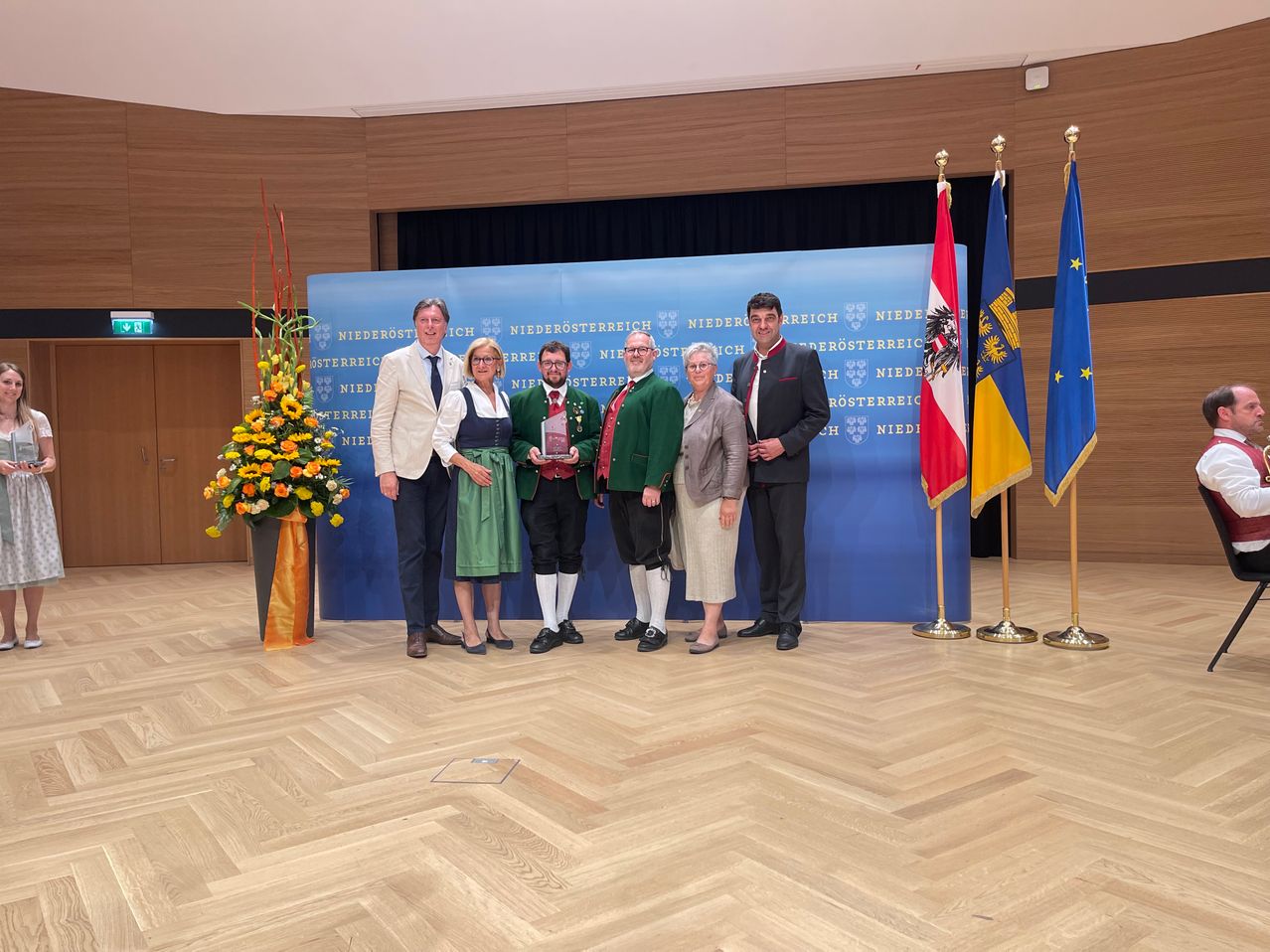 Fünf Menschen posieren für ein Foto und halten einen Pokal. Hinter ihnen steht ein blauer Banner mit weißem Text und drei Flaggen auf der rechten Seite. Der Boden ist aus Holz.