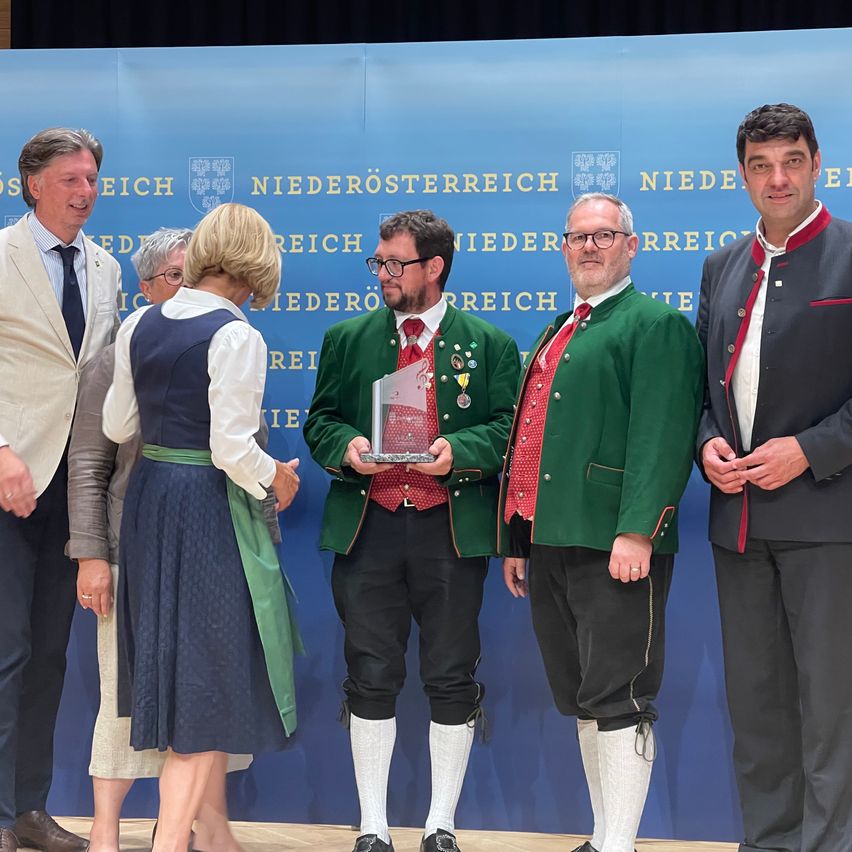 Fünf Personen stehen vor einem blauen Hintergrund und lächeln. Zwei Männer halten eine Trophäe und eine Frau steht neben ihnen. Sie tragen traditionelle bayerische Kleidung.