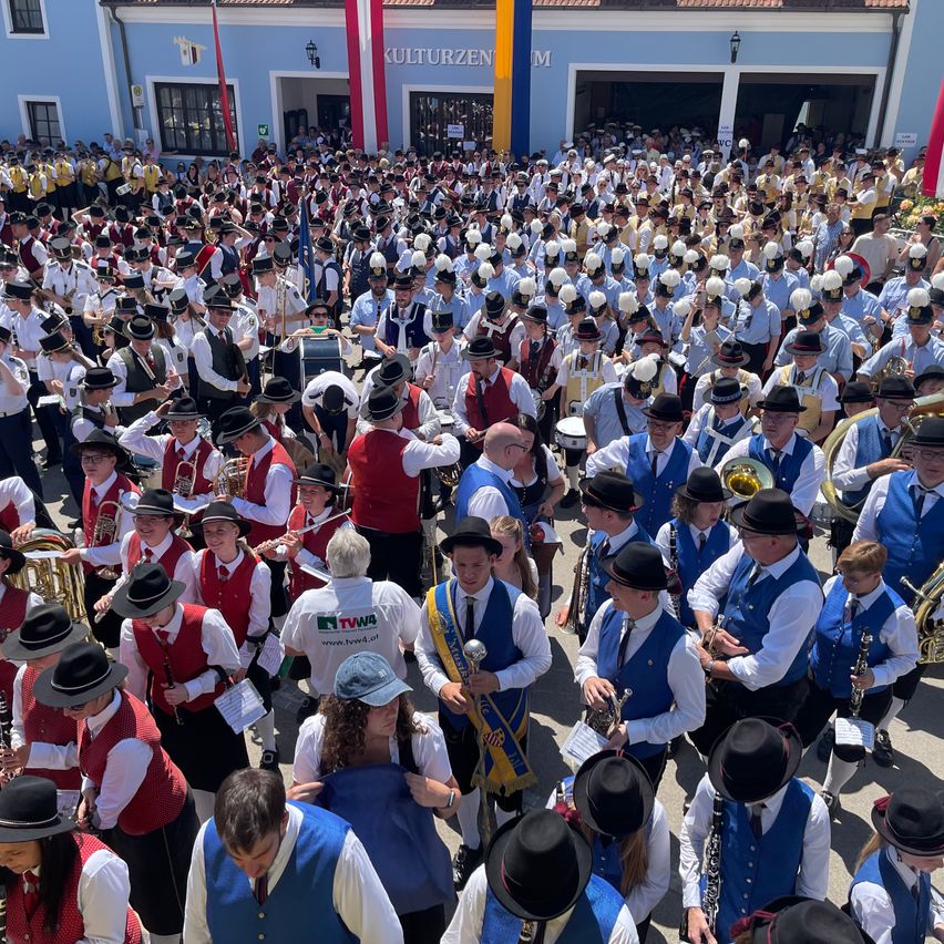 Eine große Gruppe von Musikern in traditionellen Kostümen versammelt sich im Freien und hält Instrumente. Viele tragen Hüte, Krawatten und einige haben Schärpen. Im Hintergrund befindet sich ein Gebäude mit einem Banner und Flaggen.