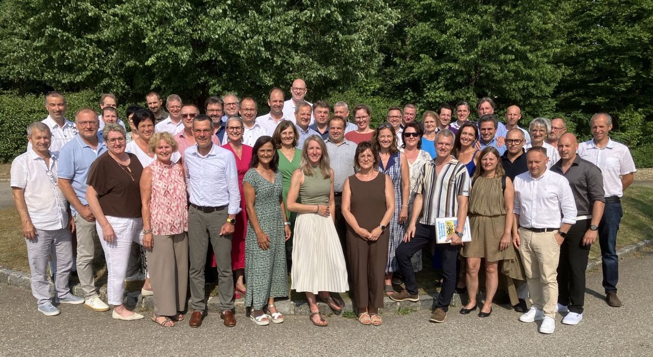 A large group of people, both men and women, are posing for a photo outdoors, smiling and standing in front of trees.