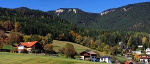 Eine ländliche Szenerie mit einem Haus im Vordergrund, grünen Feldern und Bergen im Hintergrund. Der Himmel ist klar, und es gibt mehrere Bäume und Häuser.