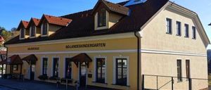 Ein gelbes Gebäude mit einem braunen Dach und mehreren Fenstern, beschriftet mit 'NOLANDENKINDERGARTEN', hat Vordächer über dem Eingang. Eine Bank und ein Zaun stehen vor dem Gebäude, und im Hintergrund sind Bäume zu sehen.