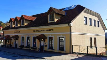 Ein gelbes Gebäude mit einem braunen Dach und mehreren Fenstern, beschriftet mit 'NOLANDENKINDERGARTEN', hat Vordächer über dem Eingang. Eine Bank und ein Zaun stehen vor dem Gebäude, und im Hintergrund sind Bäume zu sehen.