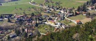 Luftaufnahme eines Dorfes in einem Tal, umgeben von grünen Feldern, Bäumen und entfernten Bergen.