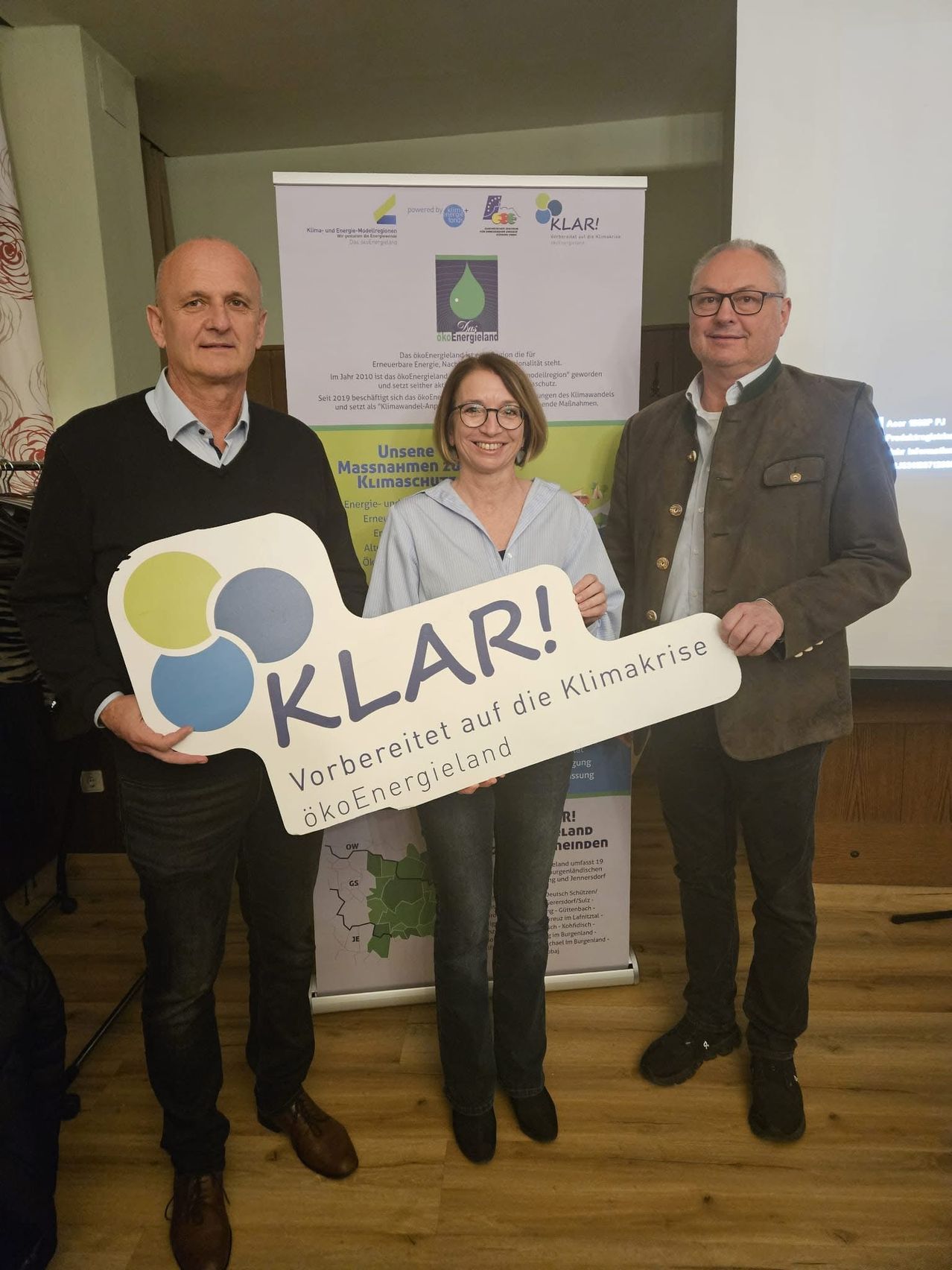 Drei Personen posieren für ein Foto mit einem Schild in einem Raum. Die Frau in der Mitte hält ein Schild mit der Aufschrift KLAR! auf weißem Hintergrund. Der Mann links und der Mann rechts lächeln. Hinter ihnen steht ein Plakat mit Text und Bildern.
