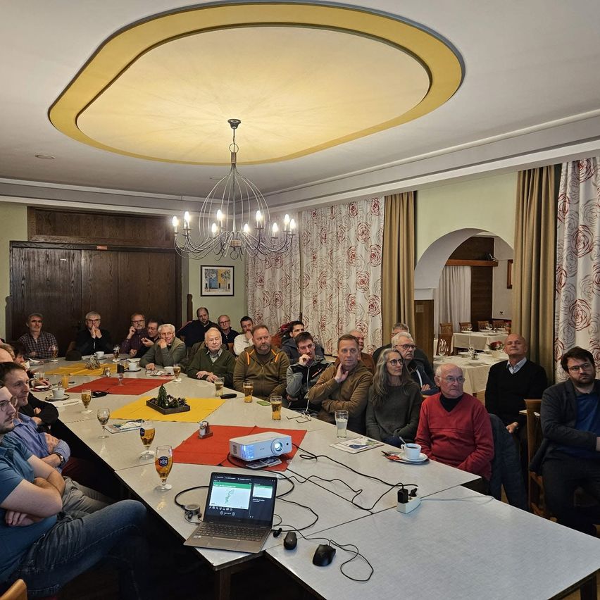 Eine Gruppe von Menschen sitzt um einen langen Tisch in einer Besprechung. Sie hören einer Präsentation zu, mit einer Projektionsleinwand und einem Laptop auf dem Tisch. Der Raum ist mit Vorhängen und einer Laterne eingerichtet.
