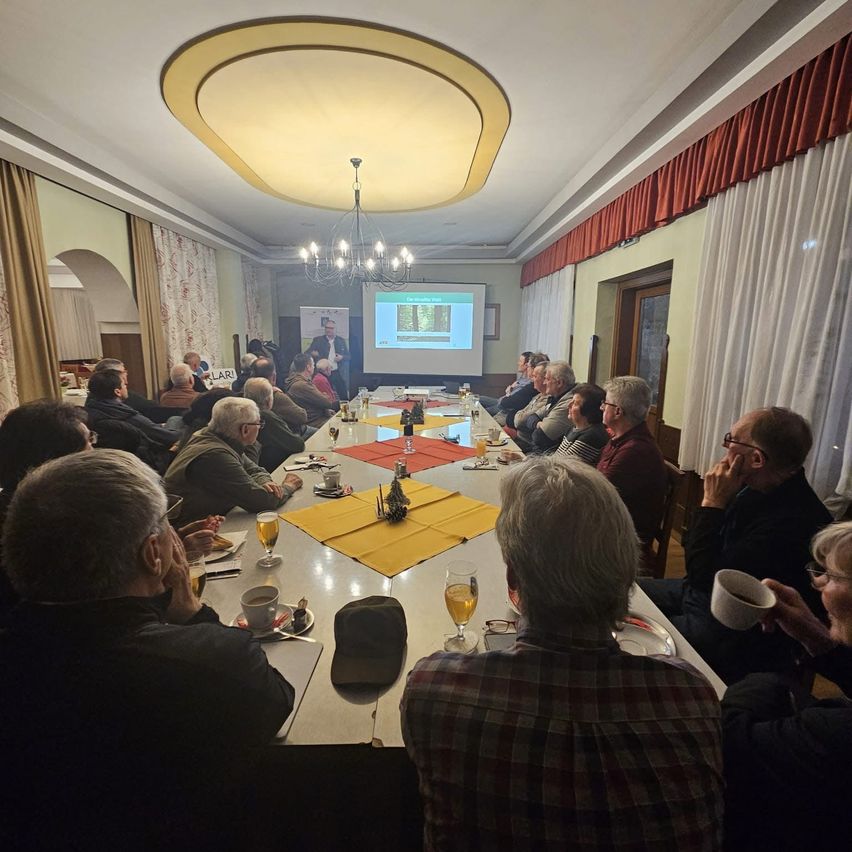 Eine Versammlung mit einem Mann, der Folien vorstellt, zu einer Gruppe von Menschen, die um einen langen Tisch herum sitzen.