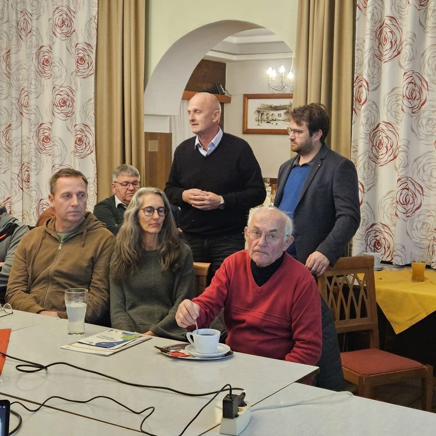 Eine Gruppe von Menschen in einem Raum, ein älterer Mann in Rot rührt Kaffee, während andere zuhören. Ein Mann in Schwarz spricht, und ein Bild hängt an der Wand.