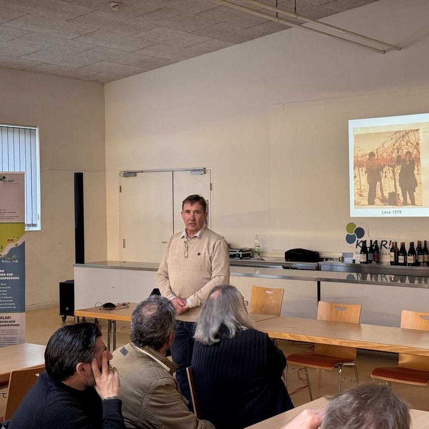 A man presents a slide showing a picture from 1978, in a room with wine bottles on the counter. Several people sit around a table, listening.