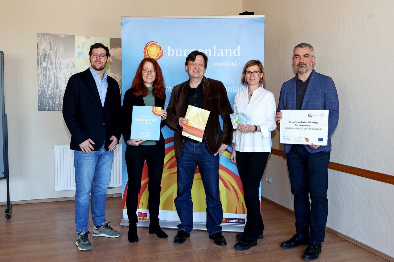 Five people stand in a room holding certificates. One is holding a certificate that reads 'Mobility for Communities'. They are smiling. Behind them is a banner with the text 'Burgenland Mobilität'.