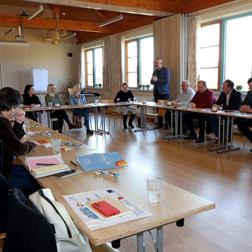 A group of people are seated around tables in a conference room. A man stands at the front, addressing the group. The room has large windows and wooden floors.