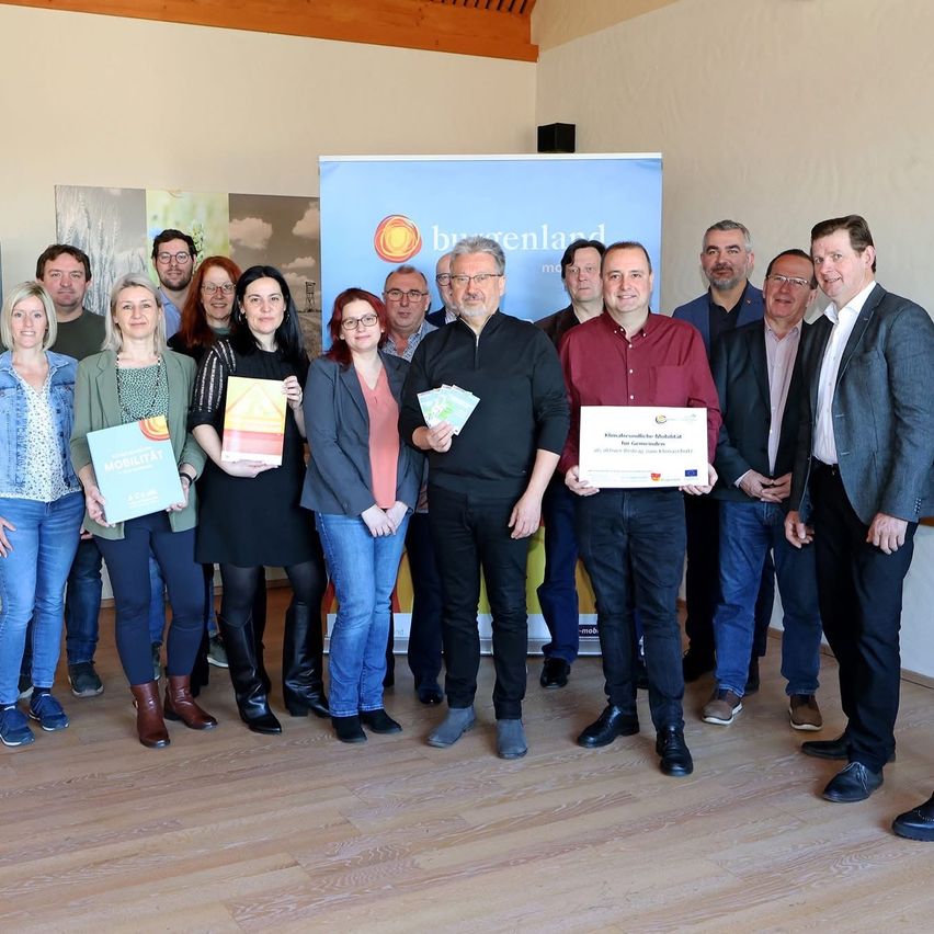 A group of people pose for a photo in a room, holding books and certificates. The banner in the background reads 'Bregenland'.