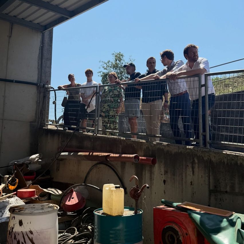 Eine Gruppe von Menschen steht auf einem Balkon und blickt auf einen verwinkelten Bereich mit Fässern und Rohren hinunter.