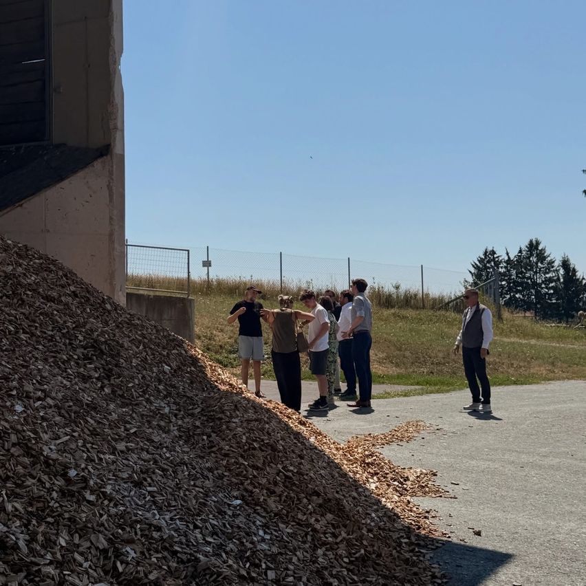 Eine Gruppe von Menschen steht neben einem Haufen Holzspäne und diskutiert möglicherweise etwas. Sie befinden sich auf einem Betonweg mit einem Zaun und Bäumen im Hintergrund.