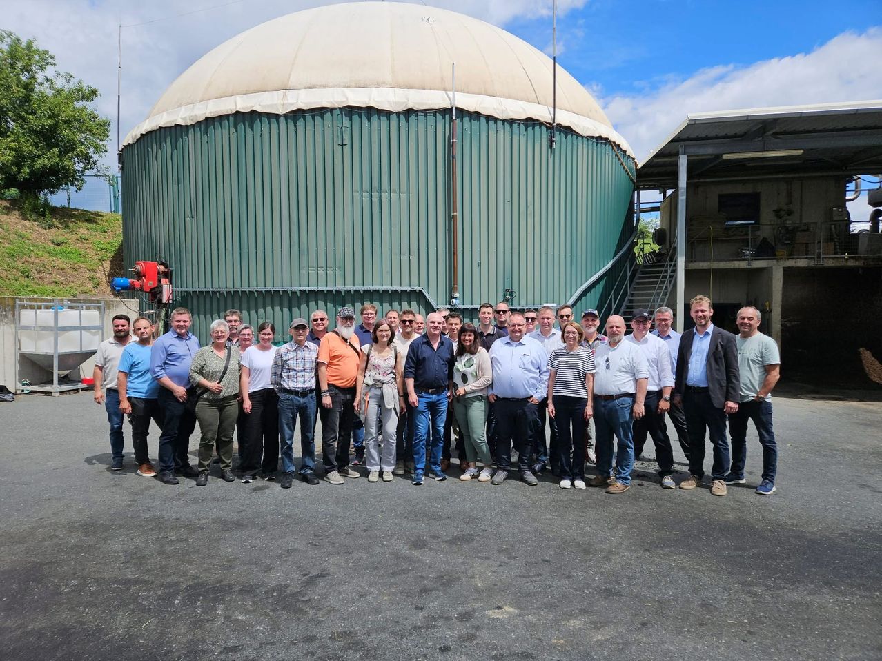 Eine Gruppe von Menschen in Freizeitkleidung posiert für ein Foto vor einem großen grünen Tank. Dahinter befinden sich Treppen, ein Gebäude und ein blauer Himmel mit Wolken.