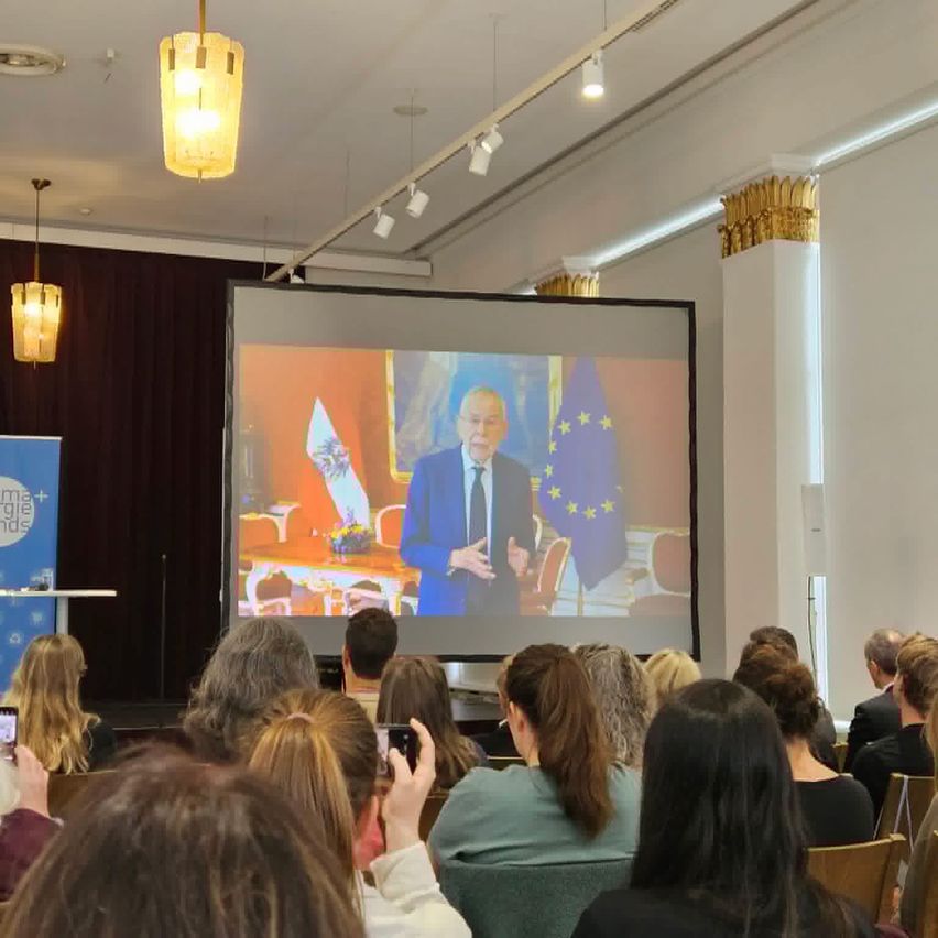 Ein Publikum sieht eine Videopräsentation auf einer großen Leinwand, mit einem Mann auf der Bühne. Flaggen sind hinter ihm angebracht. Der Raum ist gut beleuchtet mit Kronleuchtern.