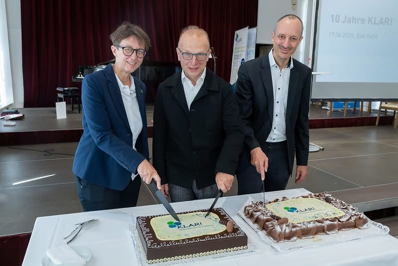 Drei Personen lächeln und posieren für ein Foto, während sie zwei Kuchen schneiden, um ein Ereignis auf einer Bühne zu feiern.