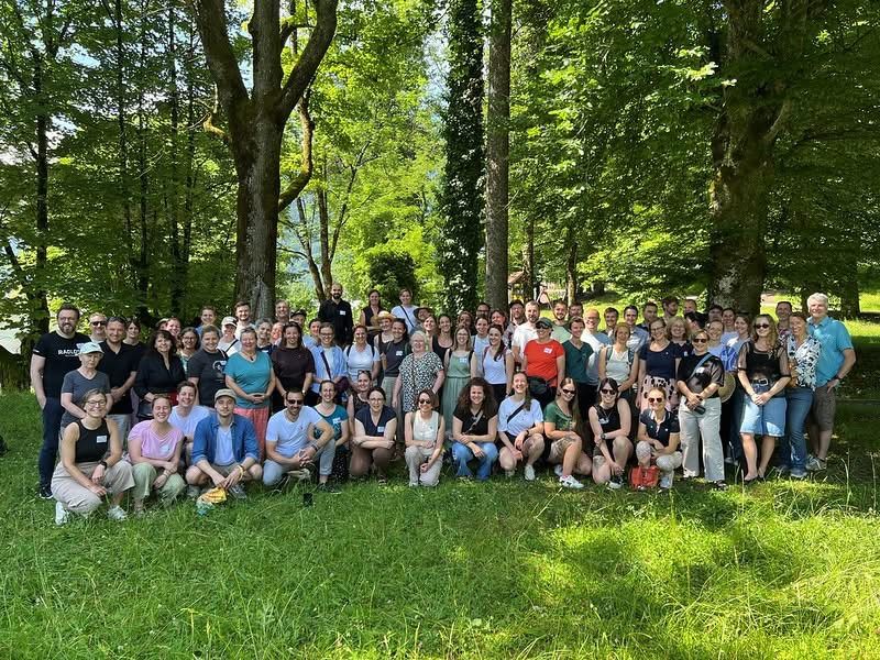 Eine große Gruppe von Menschen posiert für ein Foto in einem Wald, umgeben von hohen Bäumen und saftigem Gras.