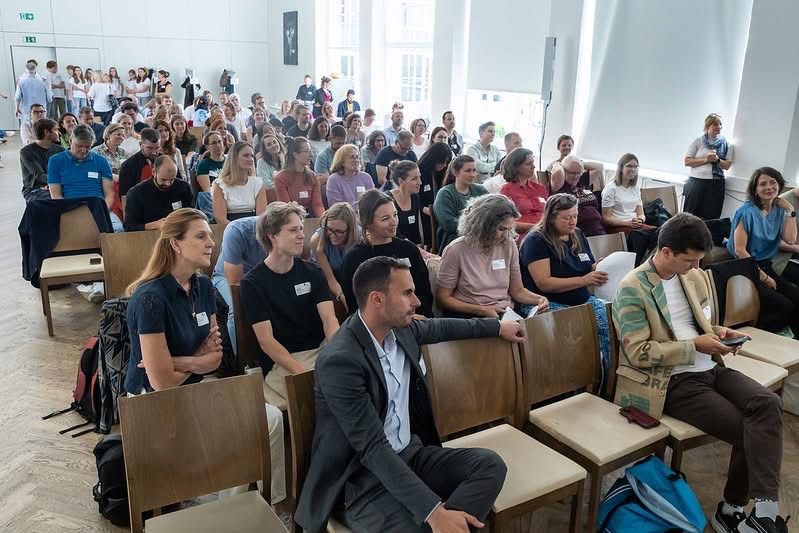 Ein großes Publikum ist in einem Hörsaal versammelt, sitzt auf Holzstühlen. Ein Mann in Anzug unterhält sich mit einer Frau in schwarzem Shirt.