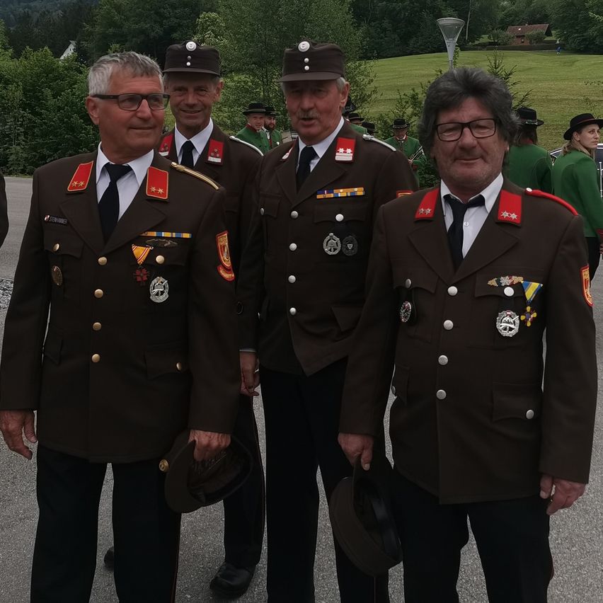 Vier Männer in Militäruniformen stehen in einem Außenbereich mit verschwommenen Menschen im Hintergrund. Sie tragen alle Mützen, Brillen und Krawatten. Sie posieren für ein Foto.