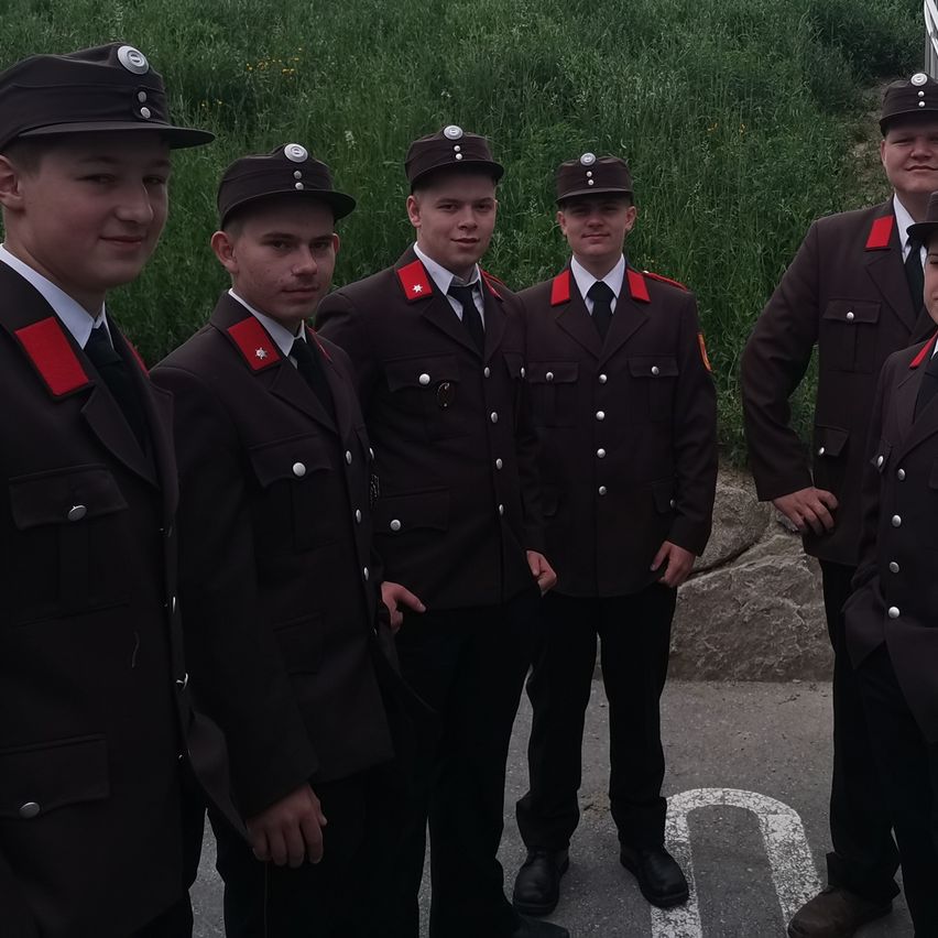 Fünf Männer in Uniformen mit roten und weißen Besätzen stehen in einer Reihe, möglicherweise für ein Foto. Sie alle lächeln, und ihre Schatten fallen auf den Boden. Dahinter befindet sich ein Grasbereich mit einer Steinmauer.