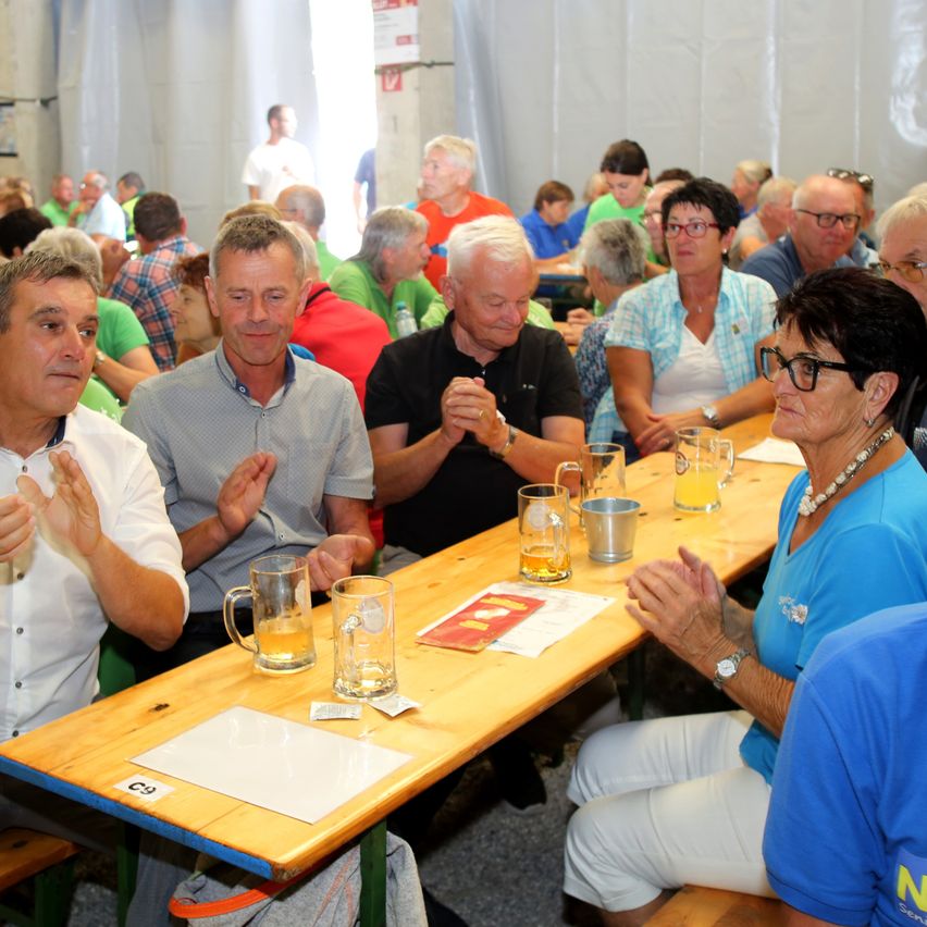 Eine Gruppe von Menschen sitzt an einem langen Tisch in einem Saal, klatscht in die Hände und lächelt. Vor jedem steht ein Glas Bier. Hinter ihnen stehen andere und unterhalten sich.