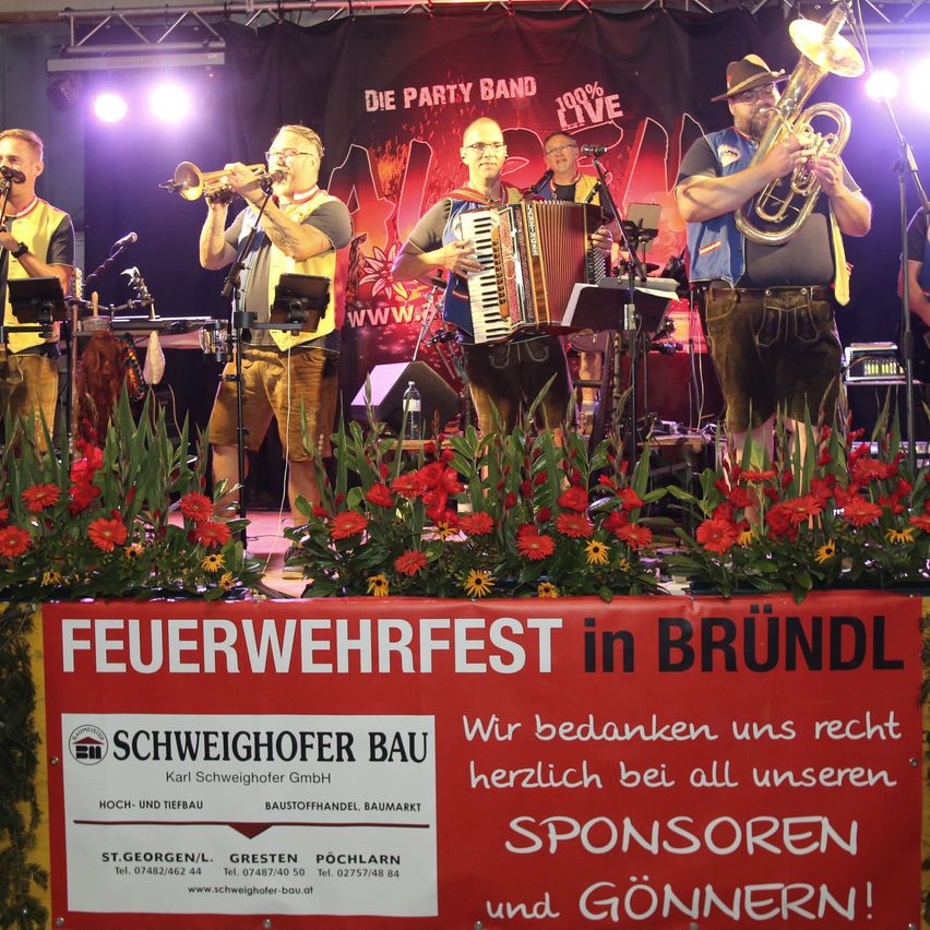Eine Band auf der Bühne spielt Musik mit Trompeten, Akkordeons und Tubas. Ein Banner trägt den Text 'Feuerwehrfest in Bründl' mit Blumen im Vordergrund.