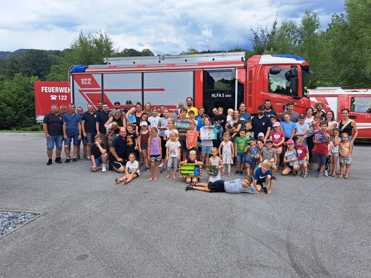 Eine große Gruppe von Menschen posiert für ein Foto vor einem roten Feuerwehrauto. Sie alle lächeln und scheinen Spaß zu haben. Einige halten Geschenke in den Händen.
