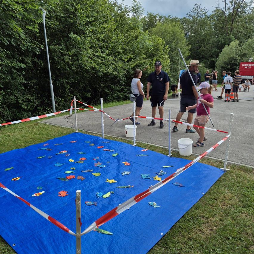 Eine Gruppe von Menschen versammelt sich um eine blaue Plane auf einem Grasfeld, auf der fischförmige Objekte verstreut sind. Zwei Personen halten Angelruten, und andere beobachten. Eimer und ein Absperrzaun sind aufgestellt.
