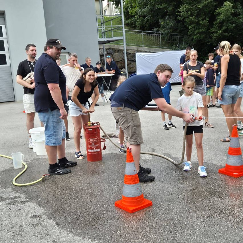 Eine Gruppe von Menschen versammelt sich um einen Mann, der einen Wasserschlauch auf ein Kind richtet. Der Bereich ist von Verkehrskegeln umgeben. Einige Leute stehen, andere sitzen an einem Tisch, und einige beobachten. Bäume und ein Gebäude sind im Hintergrund.