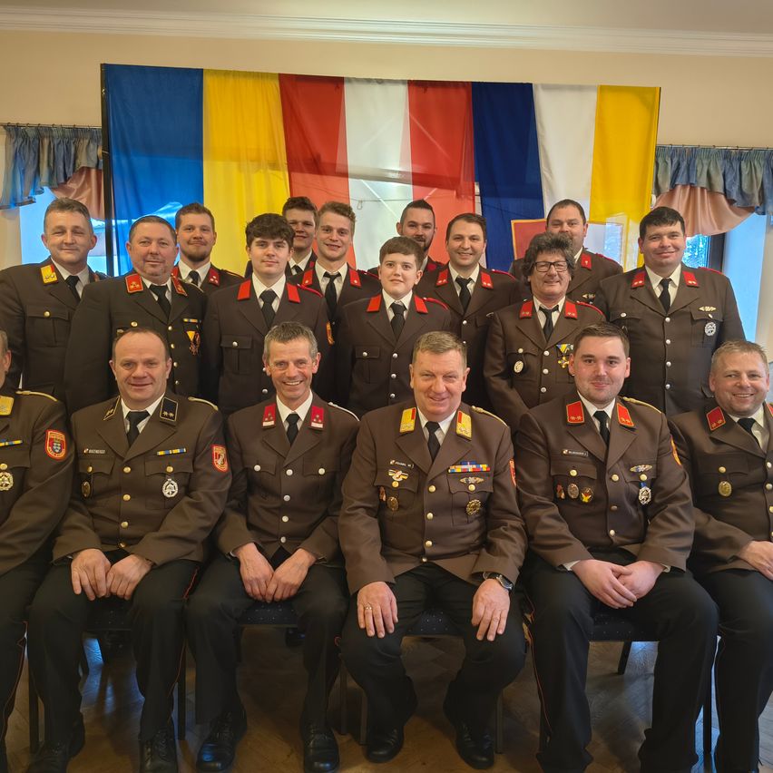 Eine Gruppe von Militärangehörigen in Uniform, sitzend und stehend, posiert für ein Foto mit Flaggen im Hintergrund.