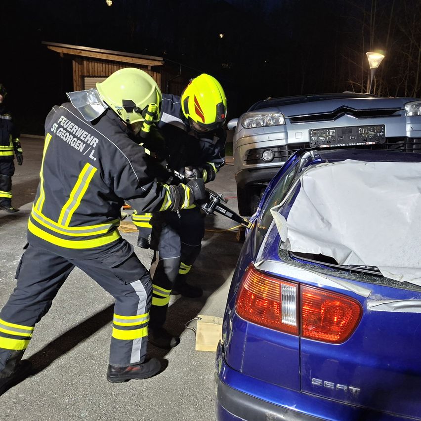 Two firefighters wearing protective gear are working on a blue car with its trunk open, while a gray car is parked nearby.