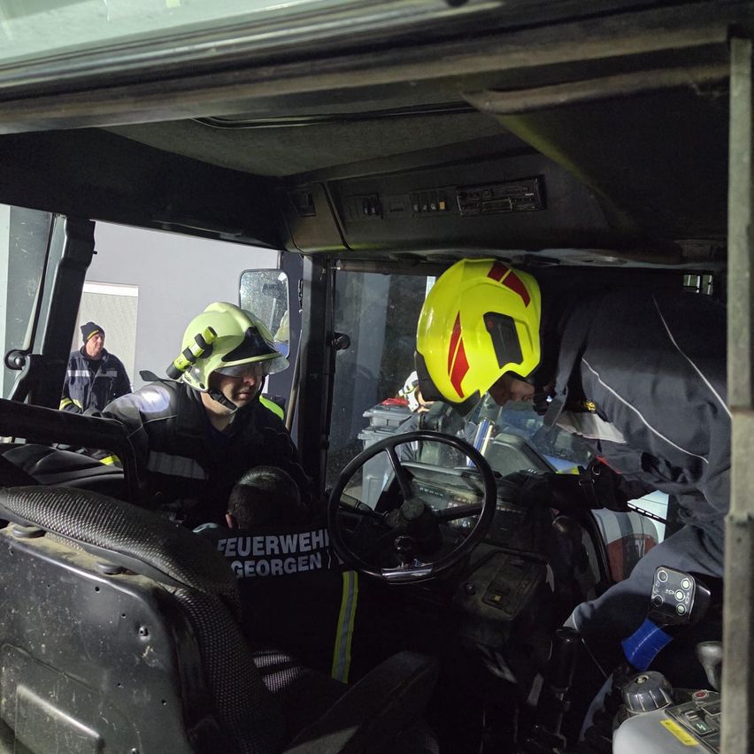A firefighter in protective gear operates a vehicle with the word 'Feuerwehr' visible. Another firefighter is seated in the front seat. They are surrounded by other firefighters.