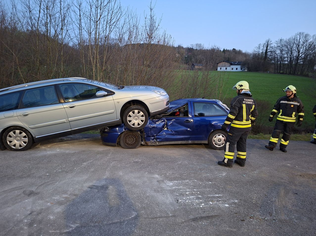 A car accident has occurred; a silver car is on top of a blue car. A firefighter stands beside the vehicles.