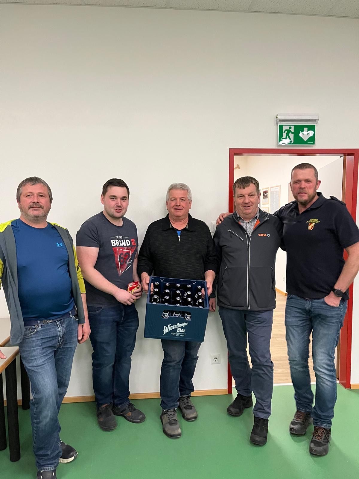 Fünf Männer posieren für ein Foto, einer hält eine Kiste Bier in einem Raum.