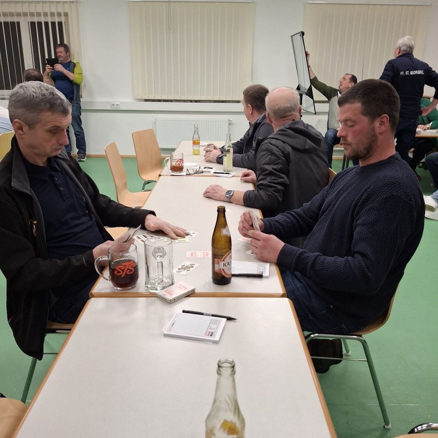 Mehrere Männer sitzen an einem langen Tisch in einem Raum, beschäftigt mit Kartenspielen, wobei einer einen Bierflasche hält und ein anderer einen Becher.