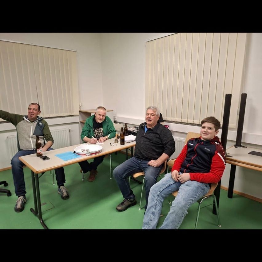 Vier Männer sitzen an einem Tisch in einem Raum, drei mit Flaschen, einer schreibt. Der Raum hat weiße Jalousien und einen grünen Boden.