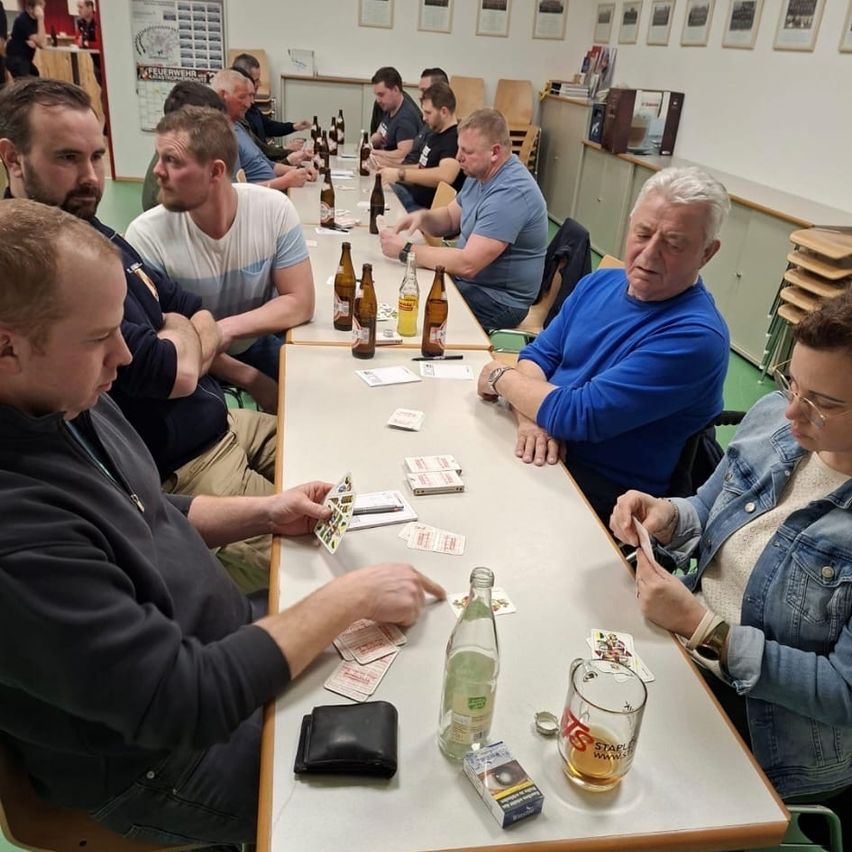 Eine Gruppe von Menschen sitzt um einen Tisch in einem Raum herum. Sie halten Karten und betrachten sie. Flaschen, Gläser und ein Geldbeutel liegen auf dem Tisch.