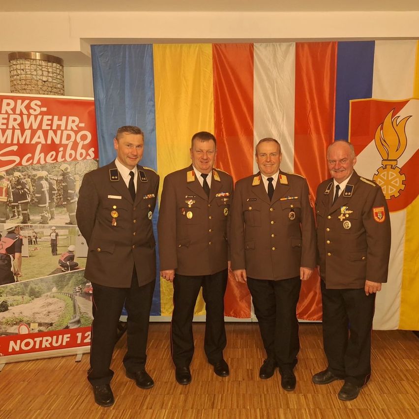 Vier Männer in Militäruniformen stehen lächelnd zusammen vor einer bunten Flagge. Sie gehören wahrscheinlich zur Armee, und einer hat ein Schild hinter sich.