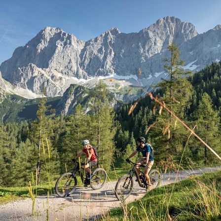 Zwei Radfahrer fahren auf einem Schotterweg durch ein üppiges grünes Tal, mit majestätischen Bergen im Hintergrund unter einem klaren blauen Himmel.