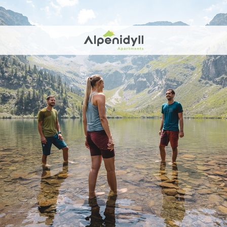 Drei Personen stehen lächelnd in einem flachen alpinen See, mit dem Logo der Alpenidyll Apartments sichtbar. Eine Bergkulisse mit grünen Bäumen und felsigem Gelände umgibt sie.