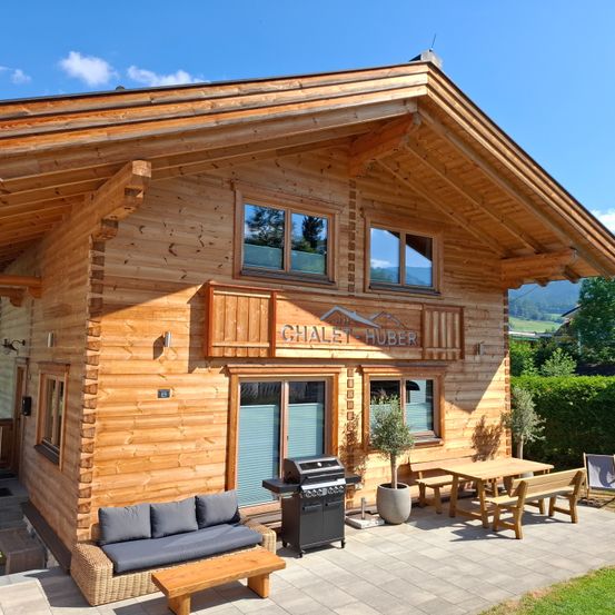 Ein Holzchalet mit geneigtem Dach, mehreren Fenstern und einer Veranda mit Sofa, Holzbank, Tisch und Grill. Es heißt Chalet Huber.