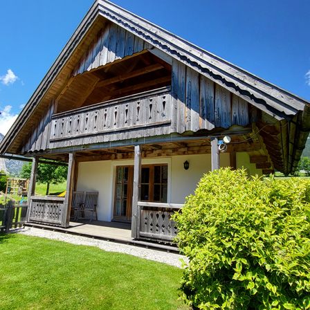 Ein Holzchalet mit Veranda, Balkon und einer Lampe, umgeben von einem grünen Rasen und einem Busch, unter einem klaren blauen Himmel.