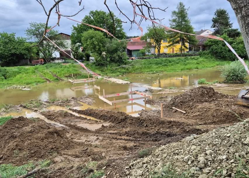 Eine Baustelle mit Holzkonstruktionen in einem überfluteten Bereich, mit Gebäuden, Bäumen und einem Feuerwehrwagen im Hintergrund.