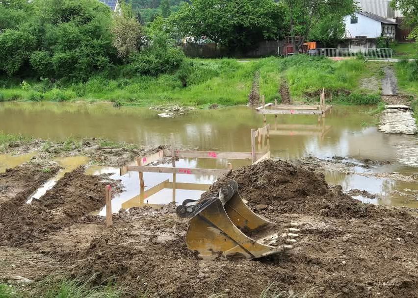 Eine Baustelle mit einem großen Bagger im Vordergrund, einem überfluteten Bereich in der Mitte und einem Haus mit Zaun im Hintergrund.